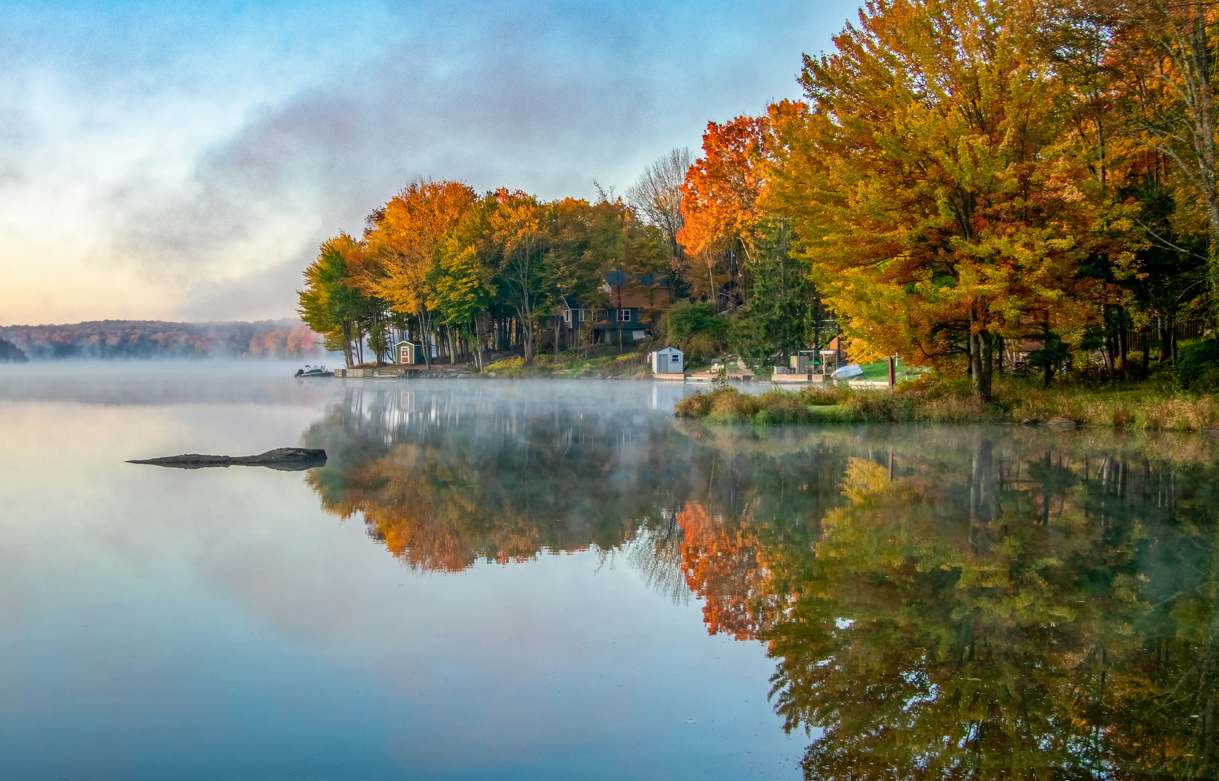 Misty autumn morning on a calm lake, with lakefront homes beneath bright fall foliage